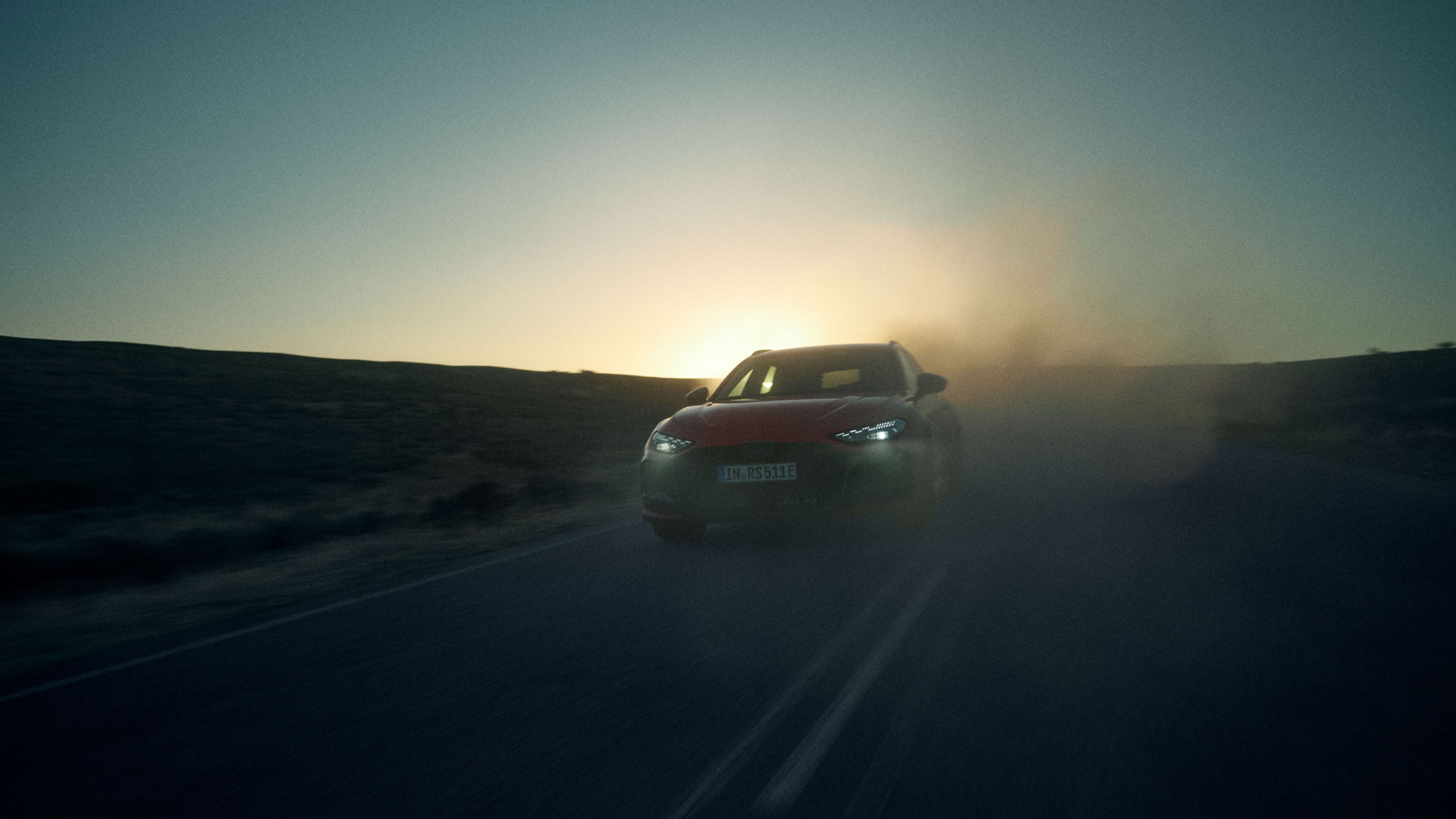 Audi RS5 vozi po prazni, podeželski cesti. Sonce je nizko na obzorju in ustvarja silhueto avtomobila. Okolica je temna in redko poseljena, cesta pa sega v daljavo.