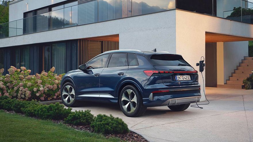 Temno moder Audi Q4 SUV e-tron je parkiran pred moderno dvonadstropno hišo z velikimi steklenimi okni. Avto je priključen na polnilno postajo ob dovozu. Okoli hiše so urejene gredice z zelenjem in cvetočimi grmovnicami.