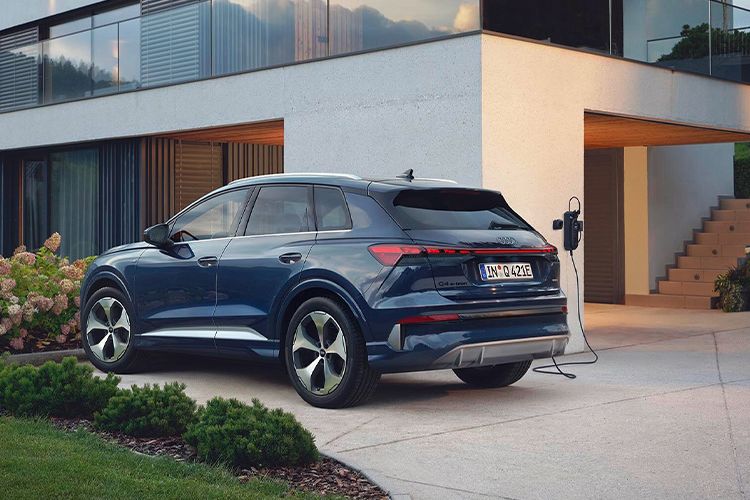 Temno moder Audi Q4 SUV e-tron je parkiran pred moderno dvonadstropno hišo z velikimi steklenimi okni. Avto je priključen na polnilno postajo ob dovozu. Okoli hiše so urejene gredice z zelenjem in cvetočimi grmovnicami.