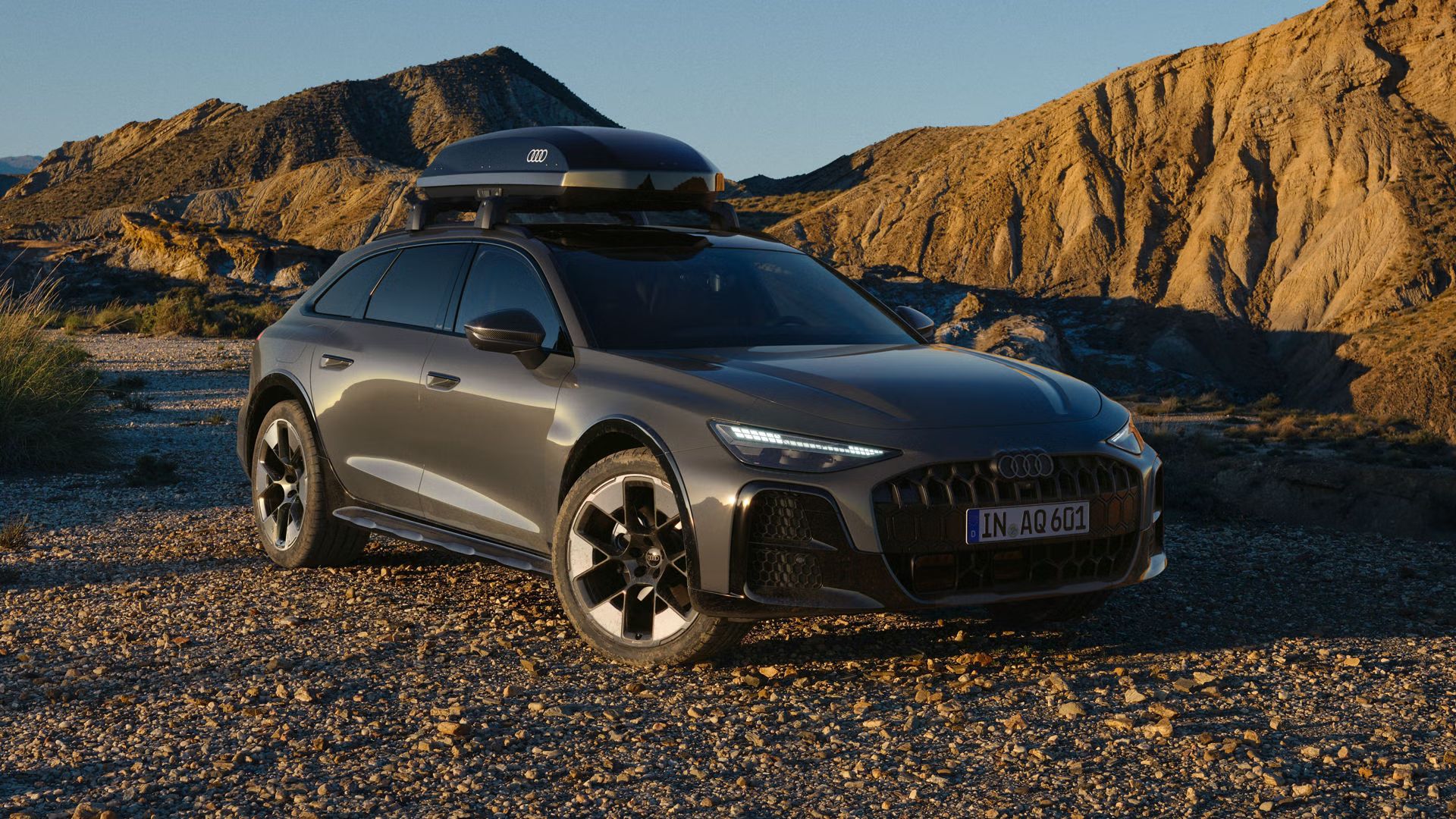 Audi A6 allroad, prikaz od spredaj, parkira na makadamski cesti. V ozadju je pusta in skalnata pokrajina, sončna svetloba s strani pa prizor razdeli na senčne in osvetljene dele.