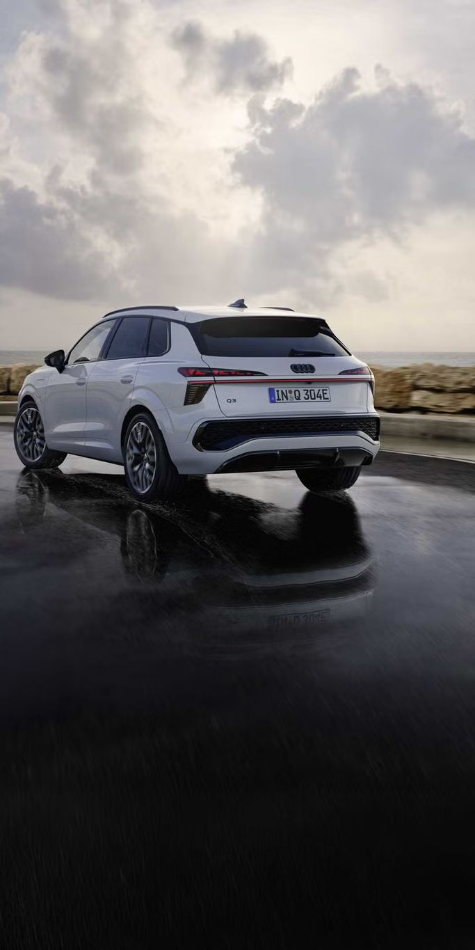 Ein weißer Q3 SUV e-hybrid von hinten fährt auf nasser Straße entlang der Küste.	