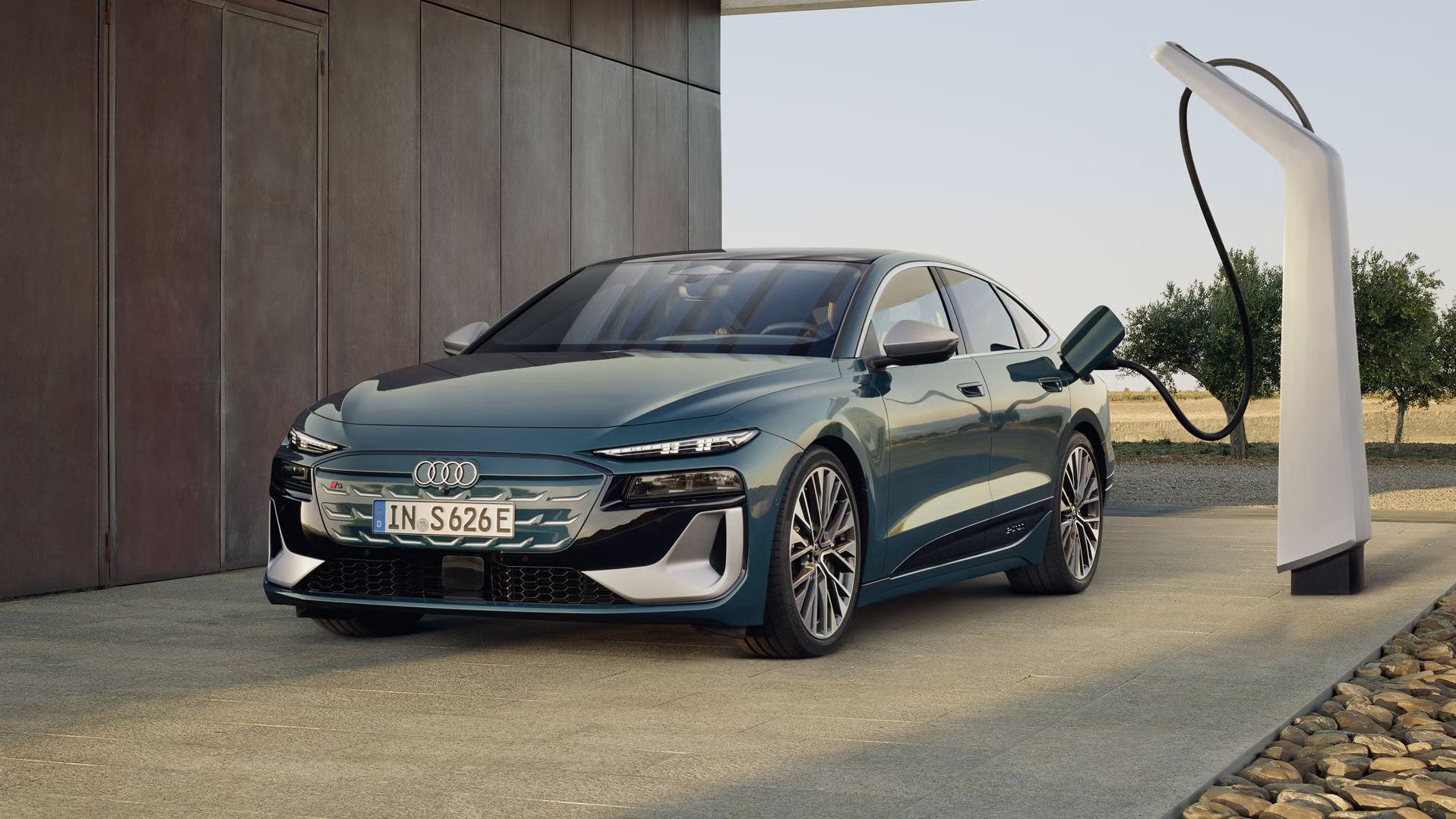 Audi S6 Sportback e-tron v kovinsko zeleni barvi, parkiran ob sodobni hiši in priključen na polnilno postajo. Avtomobil je fotografiran s sprednje-bočnega kota, z izrazito nizko in športno silhueto, velikimi platišči ter ostro oblikovanimi LED žarometi, v ozadju pa je odprta pokrajina z drevesi.