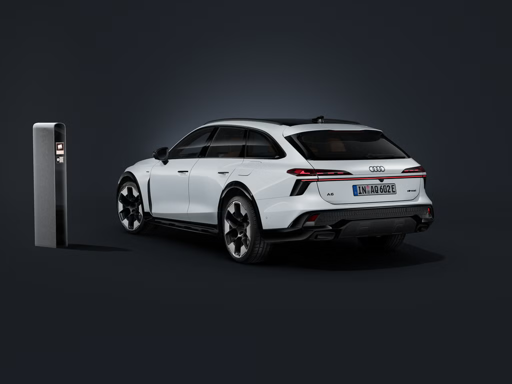 Audi A6 allroad e‑hybrid, prikazan diagonalno od zadaj na temnem ozadju, ob katerem stoji sodobna polnilna postaja.