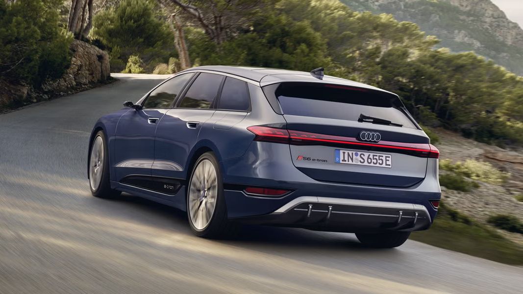 Audi S6 Sportback e-tron vozi po zaviti cesti skozi naravno okolje. Vozilo je prikazano od zadaj med vožnjo, obdano z drevesi in gričevnato pokrajino. Fotografija poudarja sodoben dizajn in električno mobilnost znamke Audi.