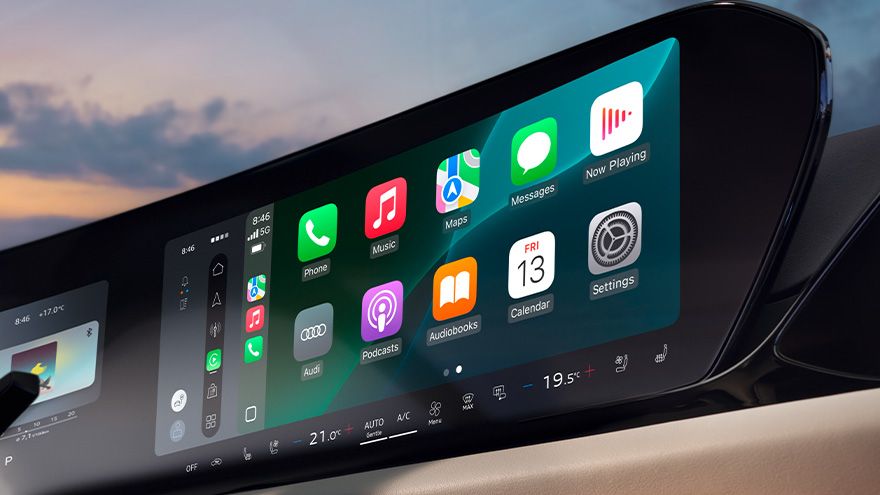 Digitalni zaslon v notranjosti avtomobila Audi A5 prikazuje širok vmesnik z ikonami aplikacij, podobnimi pametnemu telefonu (telefon, glasba, zemljevidi, sporočila, podcasti, nastavitve). Na spodnjem delu zaslona so digitalni gumbi za upravljanje klime, vključno z nastavitvijo temperature in prezračevanja. V ozadju se skozi vetrobransko steklo vidi nebo ob sončnem zahodu.