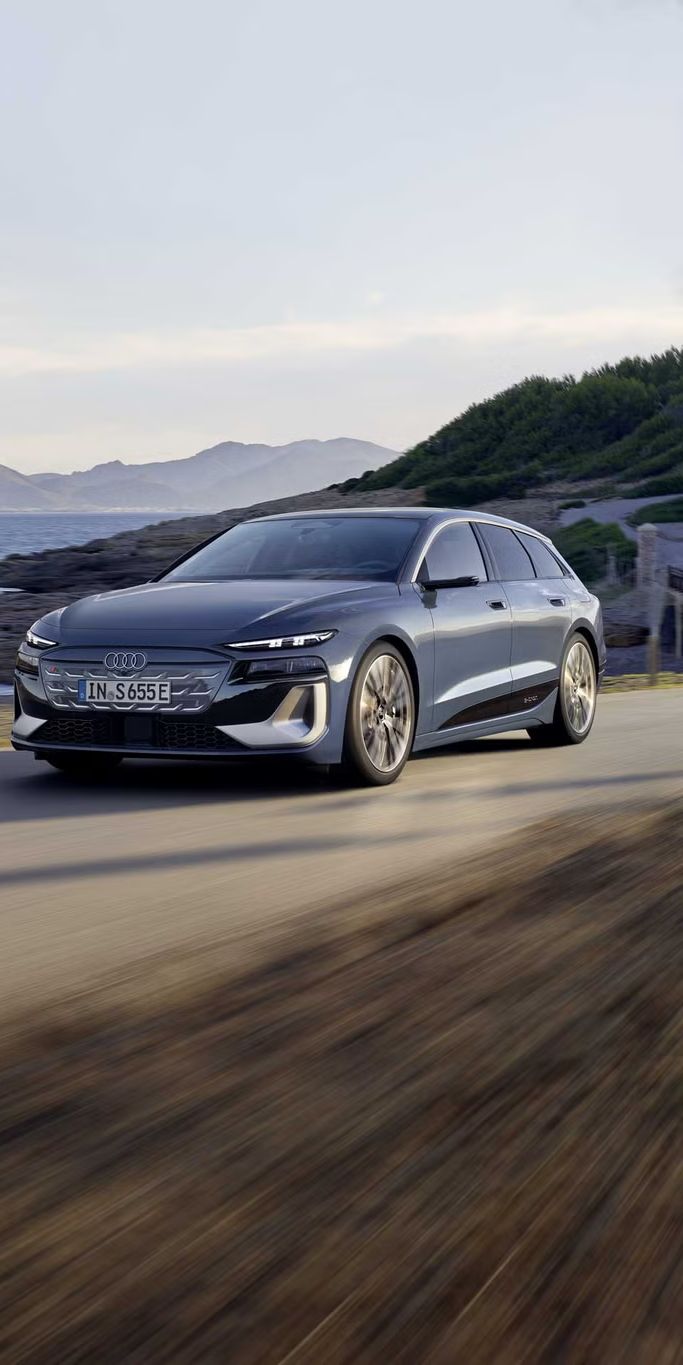 Audi S6 Avant e-tron v sivi barvi, posnet med vožnjo po obalni cesti. Avtomobil je prikazan s sprednje-bočnega kota, z razpotegnjenim karavanskim zadkom, v ozadju pa se vidijo morje, skalnata obala in hribi, kar poudarja občutek hitrosti in potovanja.