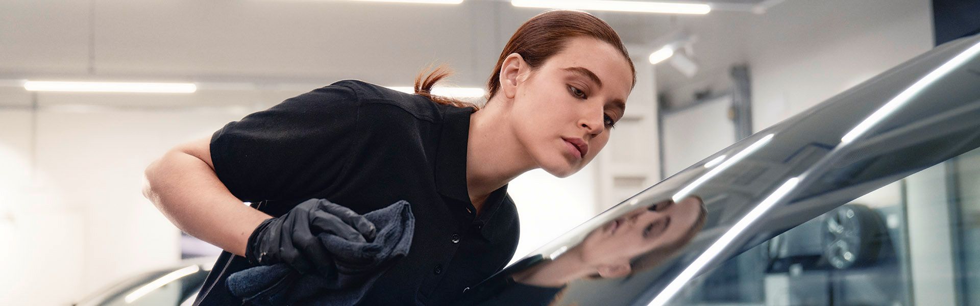 Oseba v črni majici in rokavicah čisti steklo avtomobila z mehko krpo.