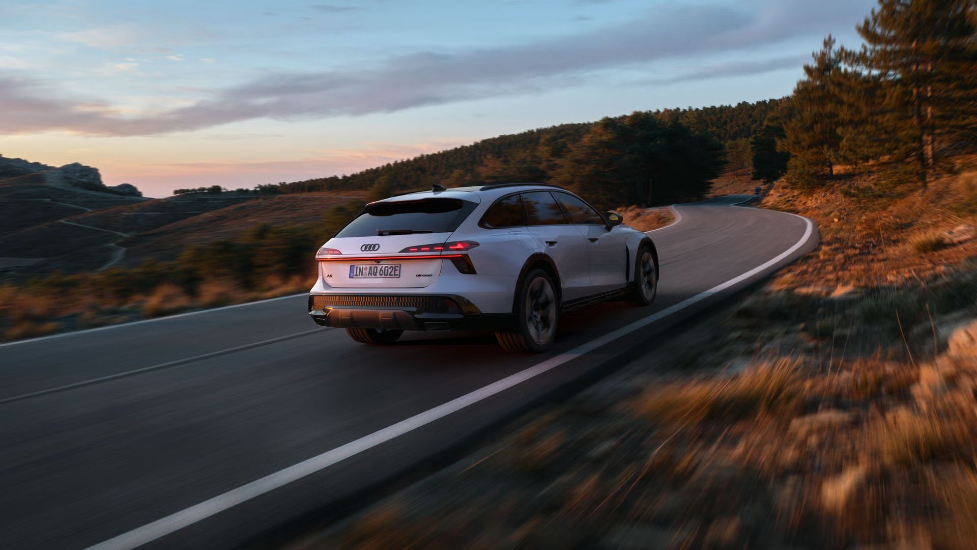 Audi A6 allroad e‑hybrid, prikazan diagonalno od zadaj pri polni hitrosti na gorski cesti; v ozadju se vidi del gozda.