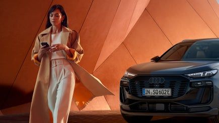 E-mobilnost v vsakdanjem življenju. Eleganten Audi SQ6 v temni barvi je parkiran pred sodobno, oranžno arhitekturno kuliso z ostrimi linijami. Ob njem hodi ženska v dolgih, svetlih hlačah in plašču, gleda v telefon in deluje samozavestno. Scena daje vtis urbanega, modernega življenjskega sloga.