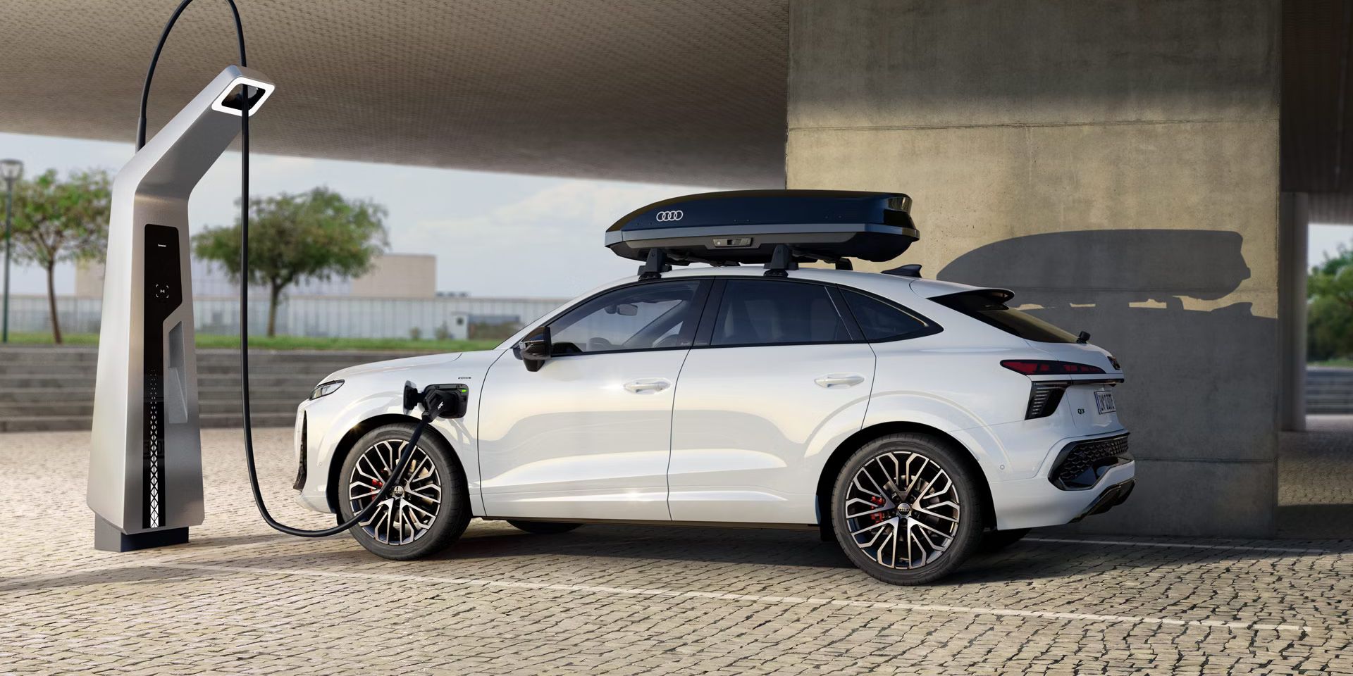 Audi Q3 SUV Sportback s strani na parkirišču med polnjenjem. Poleg tega je na belem avtomobilu vidna strešna škatla.