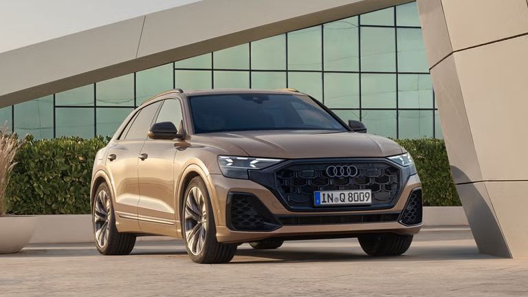 Audi Q8 SUV, prikazan v diagonalnem pogledu od spredaj, je parkiran pred sodobno arhitekturo. Urejena živa meja ustvarja eleganten prehod med vozilom in popolnoma zastekljeno stavbo v ozadju.