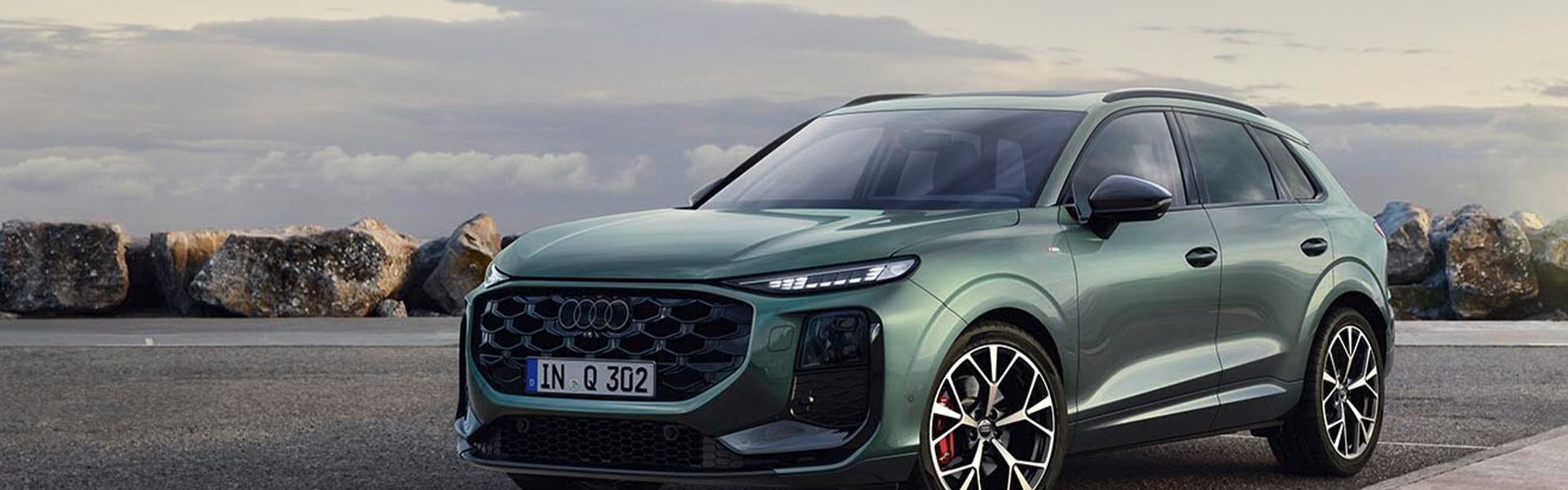 Prikaz stranskega dela zelenega vozila Audi Q3 SUV, parkiranega na parkirišču pred kamnitim zidom ob obali.