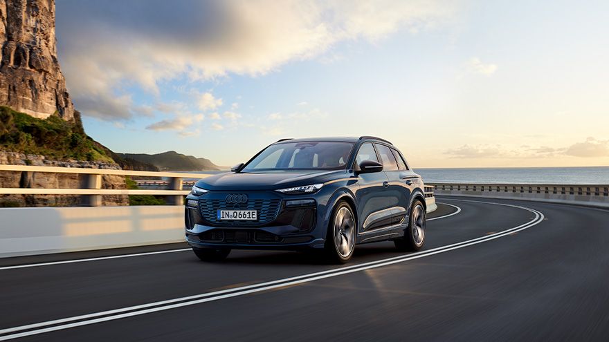 Temno moder sodoben Audi Q6 e-tron vozi po široki, ovinkasti obalni cesti ob sončnem zahodu. Na levi strani so visoke skalnate pečine, v ozadju pa se vidi morje in svetlo nebo z oblaki. Cesta je prazna in daje občutek mirne, dinamične vožnje.