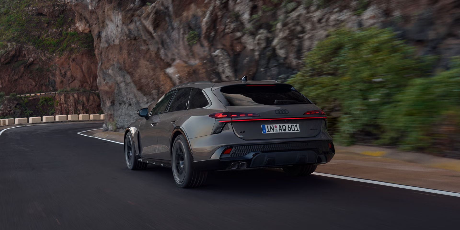 Audi A6 allraod, diagonalni pogled od zadaj, se pelje po gorski cesti. Na desni se dvigajo strme skalne stene.