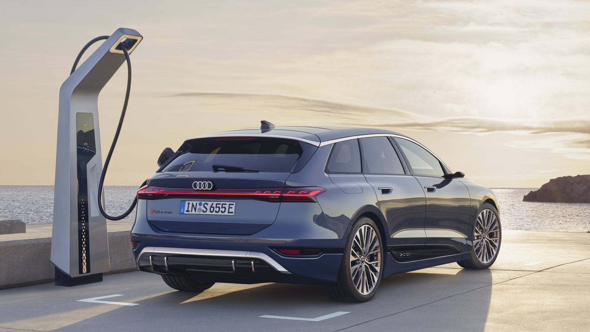 Električni Audi S6 SUV je parkiran ob polnilni postaji in priključen na polnjenje. Vozilo je prikazano od zadaj ob obalni cesti, v ozadju pa sta morje in večerno nebo. Fotografija poudarja sodoben dizajn in električno mobilnost znamke Audi.