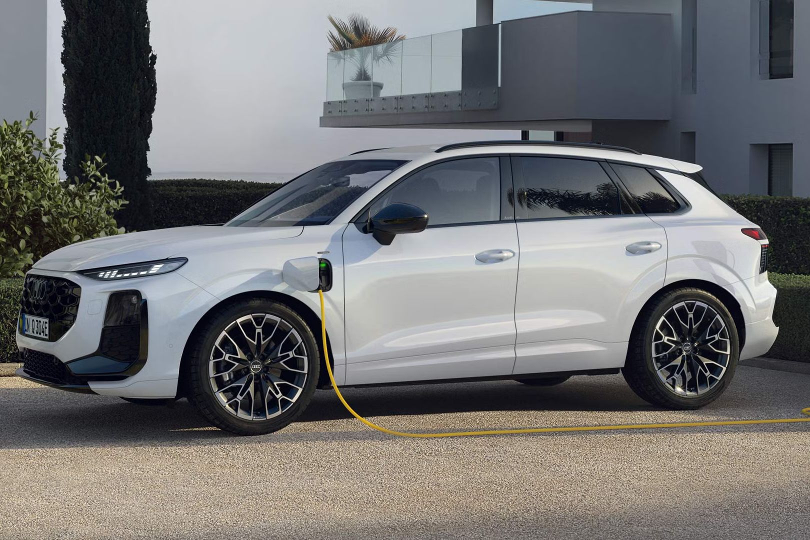 Seitenprofil eines weißen Q3 SUV e-hybrid, der auf dem Parkplatz vor einem modernen Haus lädt.