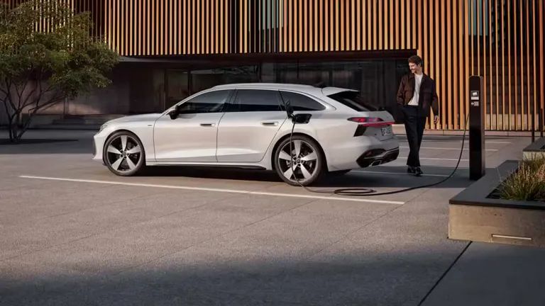 Moški stoji poleg parkiranega belega A6 Avant e-hybrid pred sodobno stavbo. Avtomobil je priključen na polnilnico, stavba pa ima lesene elemente na fasadi.  