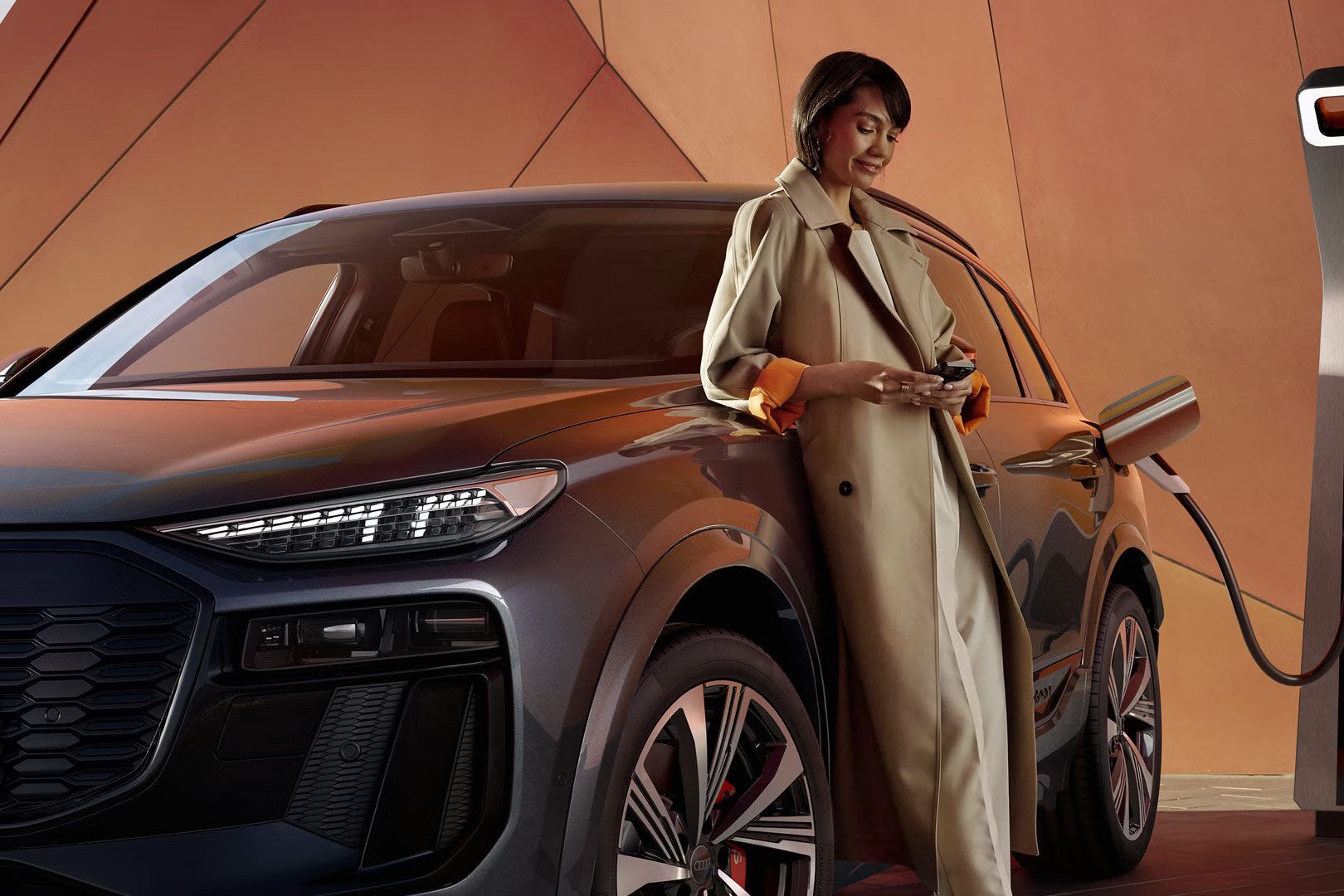 Vozilo Audi SQ6 je priključeno na električno polnilnico, nanj je naslonjena ženska, ki brska po telefonu.