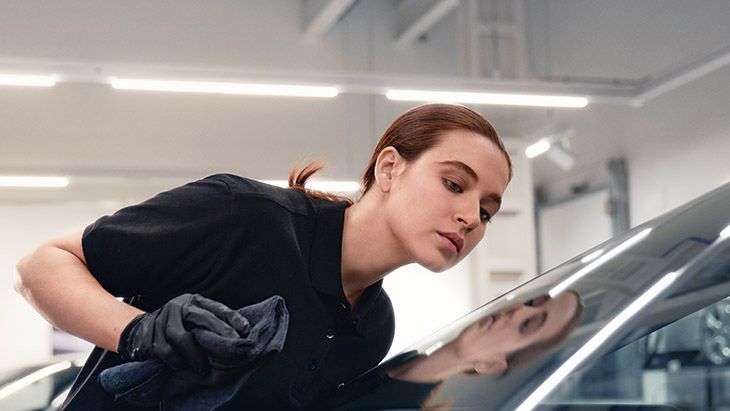 Oseba v enobarvnem oblačilu s krpo čisti prednje steklo avtomobila. Delavnica z močno osvetlitvijo. Poudarek je na čistoči in vzdrževanju avtomobila.