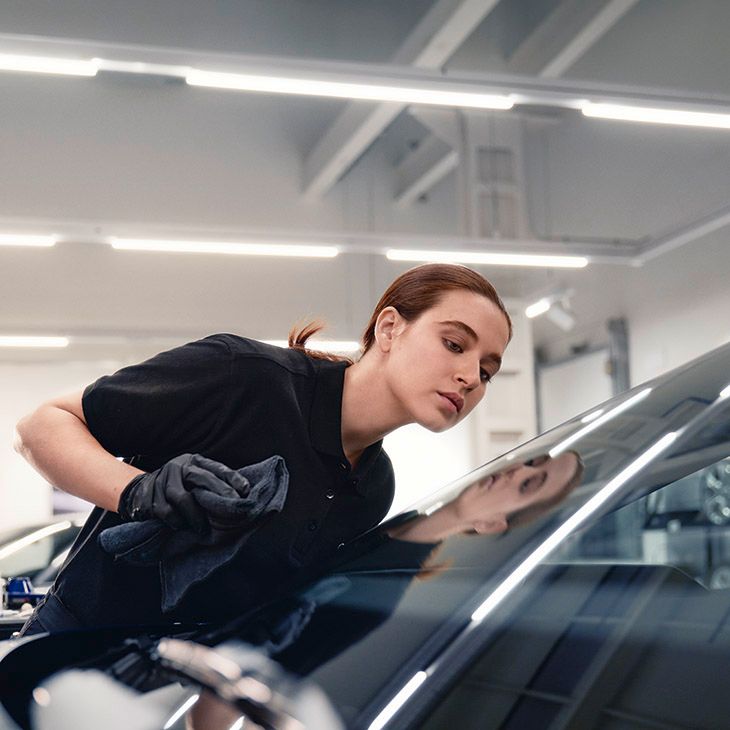 Oseba v enobarvnem oblačilu s krpo čisti prednje steklo avtomobila. Delavnica z močno osvetlitvijo. Poudarek je na čistoči in vzdrževanju avtomobila.