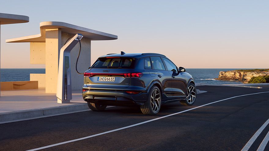 Temno moder Audi Q6 e-tron je parkiran ob polnilni postaji ob obalni cesti. Polnilni kabel je priključen v vozilo, v ozadju pa se vidita mirno morje in skalnata obala ob sončnem zahodu. Celotna scena deluje sodobno in umirjeno.