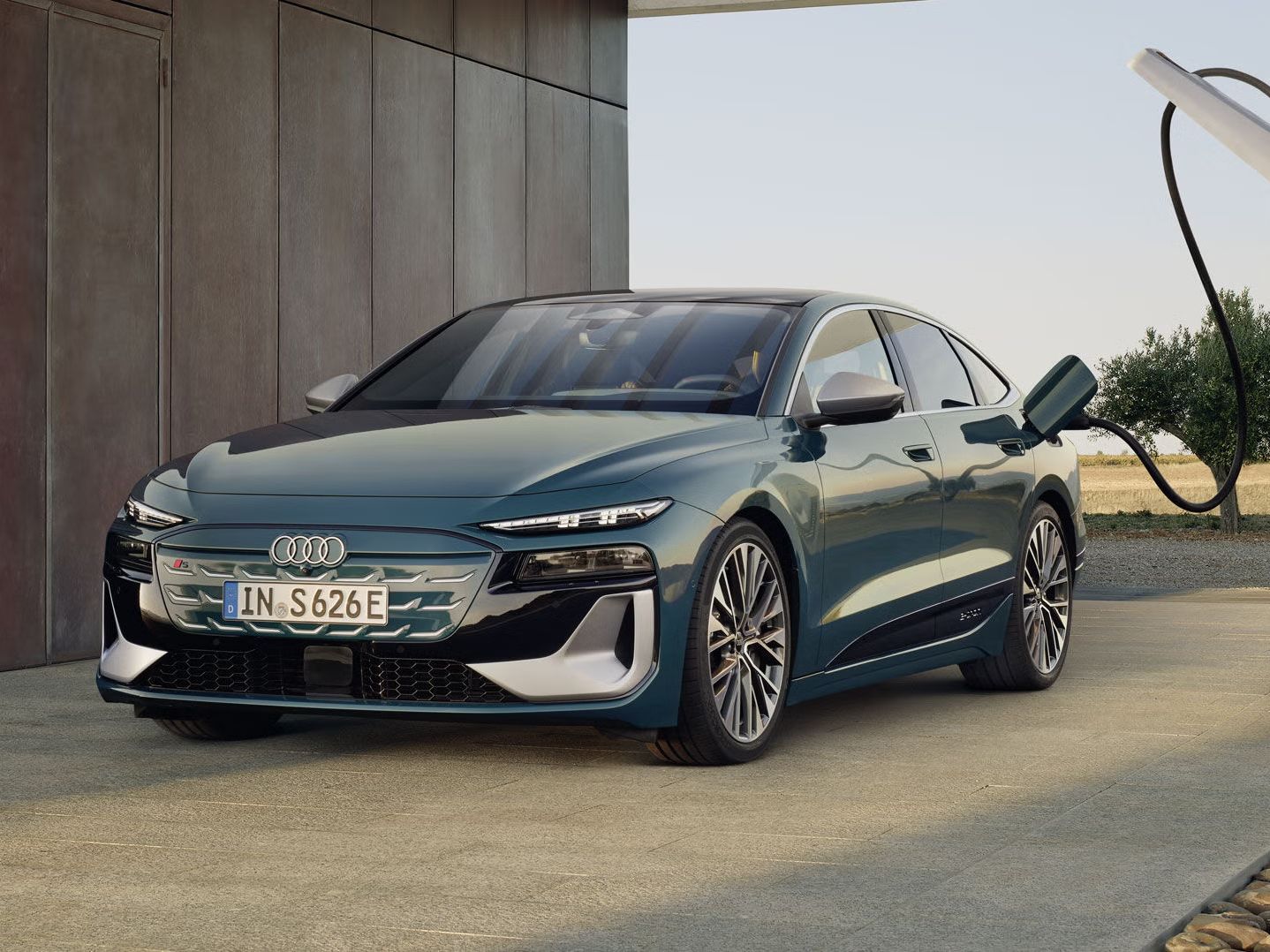 Audi S6 Sportback e-tron v kovinsko zeleni barvi, parkiran ob sodobni hiši in priključen na polnilno postajo. Avtomobil je fotografiran s sprednje-bočnega kota, z izrazito nizko in športno silhueto, velikimi platišči ter ostro oblikovanimi LED žarometi, v ozadju pa je odprta pokrajina z drevesi.