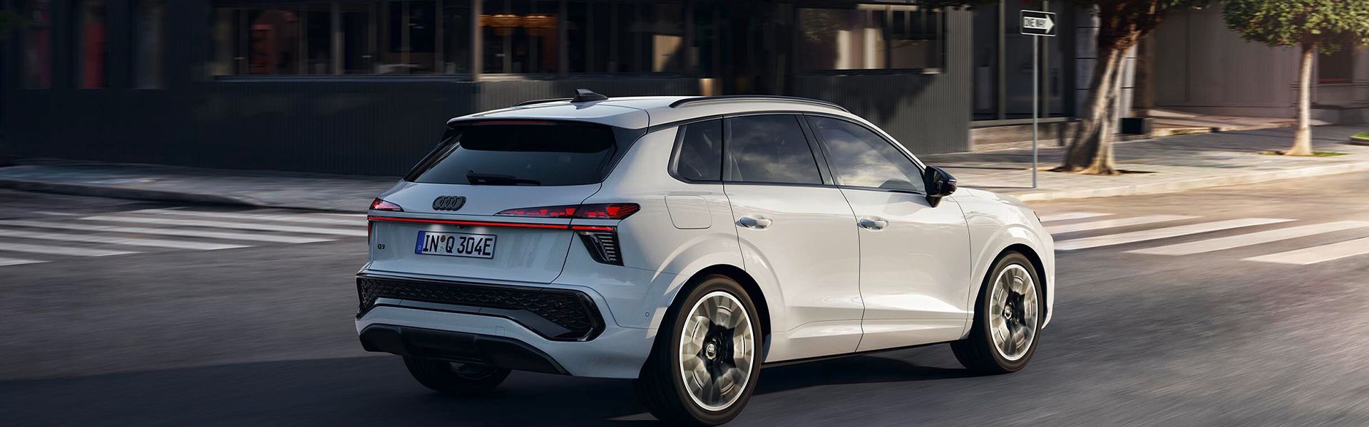 Audi Q3 SUV e‑hybrid se pelje po mestni ulici. Zadnji stranski pogled na vozilo v beli barvi.