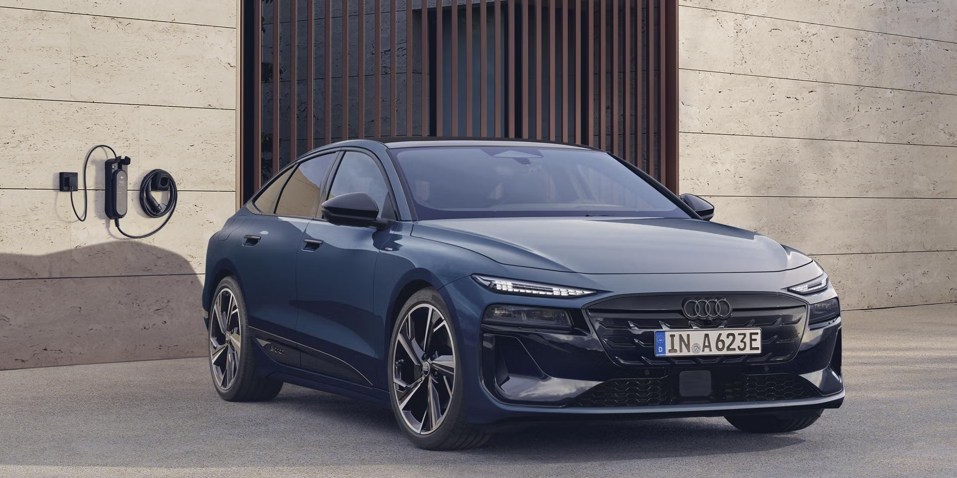 Audi S6 Sportback e-tron je parkiran pred moderno stavbo in prikazan s sprednje strani. Ob vozilu je na steni nameščena polnilna enota za domače polnjenje. Fotografija poudarja sodoben dizajn in vsakodnevno električno mobilnost znamke Audi.