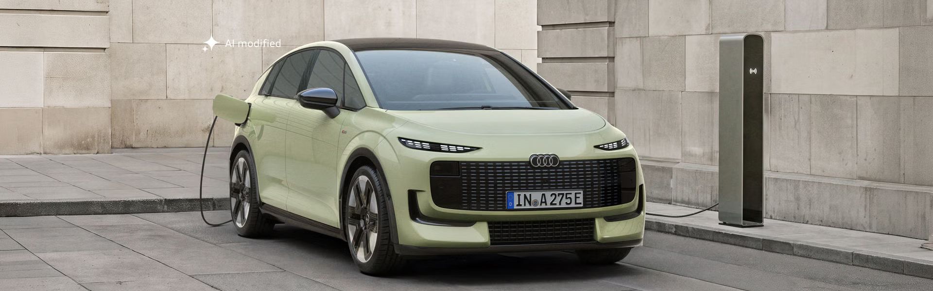Audi A2 e-tron, fotografiran s sprednje strani, je parkiran pred moderno stavbo. Vozilo je priključeno na polnilno postajo.