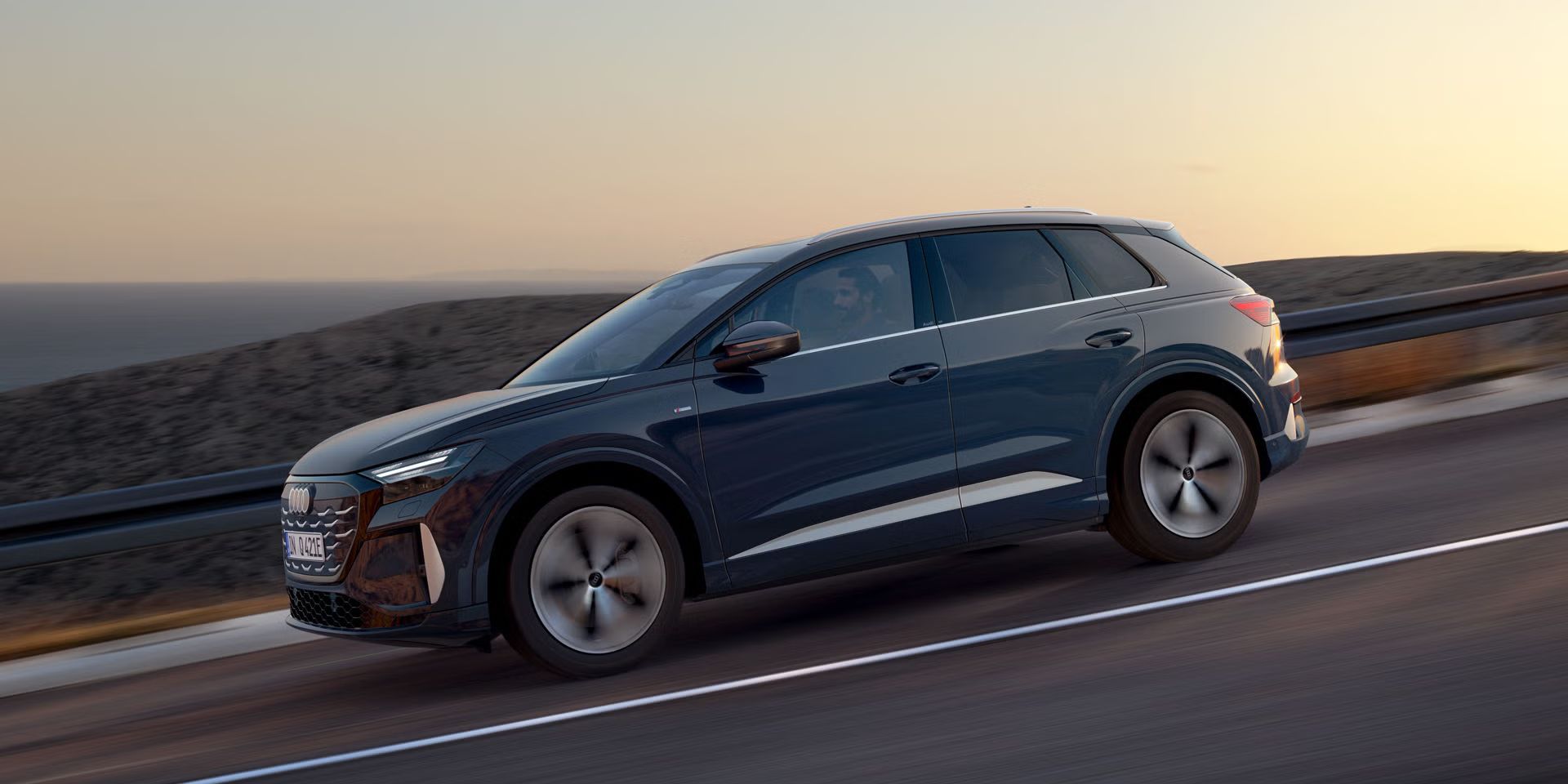 Blick von der Seite auf den Q4 SUV e-tron bei der Fahrt auf abschüssiger Straße im dämmrigen Licht. Im Hintergrund das Meer.