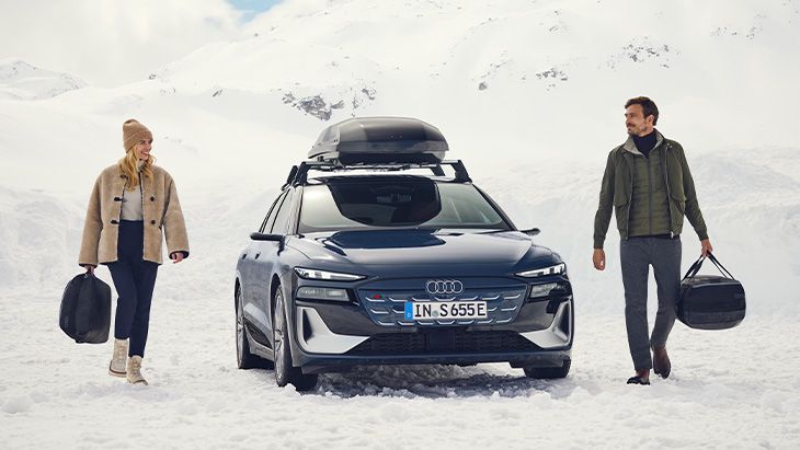 Audi A6 Avant s strešnim kovčkom, parkiran v zasneženem gorskem okolju.  Ob vozilu hodita ženska in moški, vsak z torbo v roki, oblečena v zimska oblačila.  Prizor prikazuje vozilo v zimskih razmerah ter njegovo uporabo na poti v naravnem okolju.