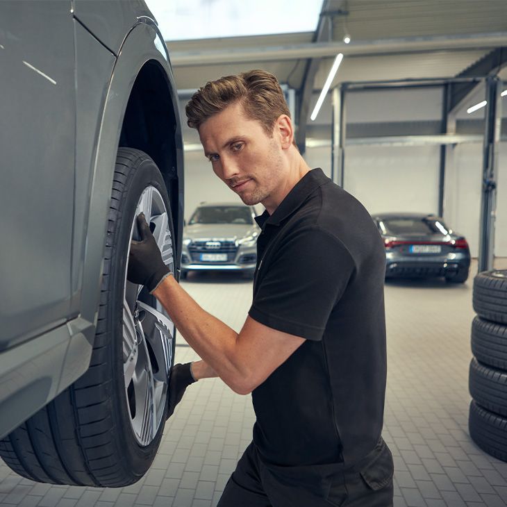 Strokovna oseba v delavnici preverja pnevmatiko vozila, dvignjenega na dvigalki. V ozadju so vidna druga parkirana vozila. Na desni strani je zložen kup štirih pnevmatik.