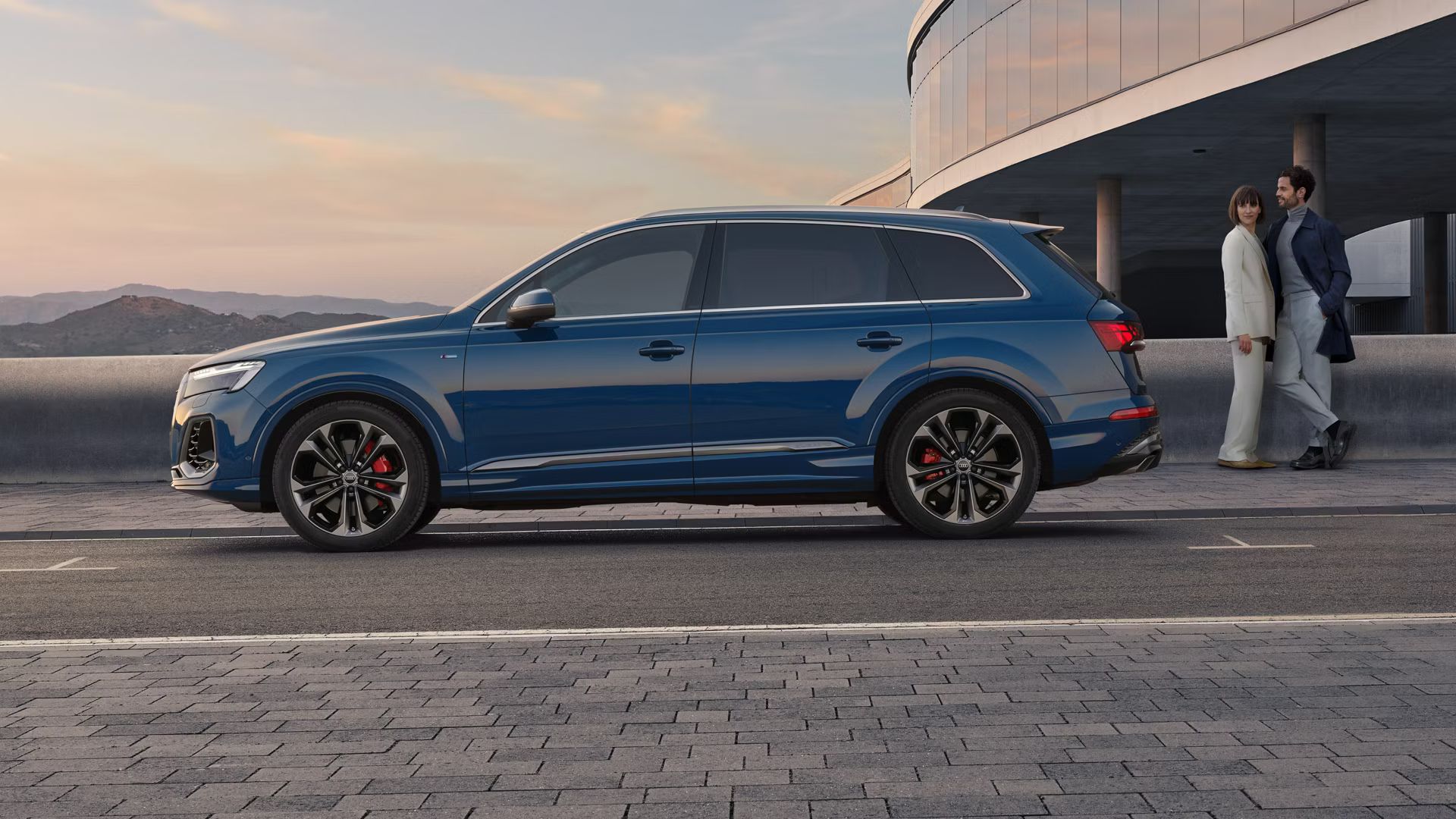Seitenansicht eines blauen Audi Q7, eine Frau und ein Mann stehen hinter dem Fahrzeug.