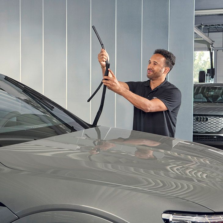 Moški z zabrisanim obrazom stoji poleg avtomobila in menja brisalec na vetrobranskem steklu. Več avtomobilov parkiranih v ozadju.