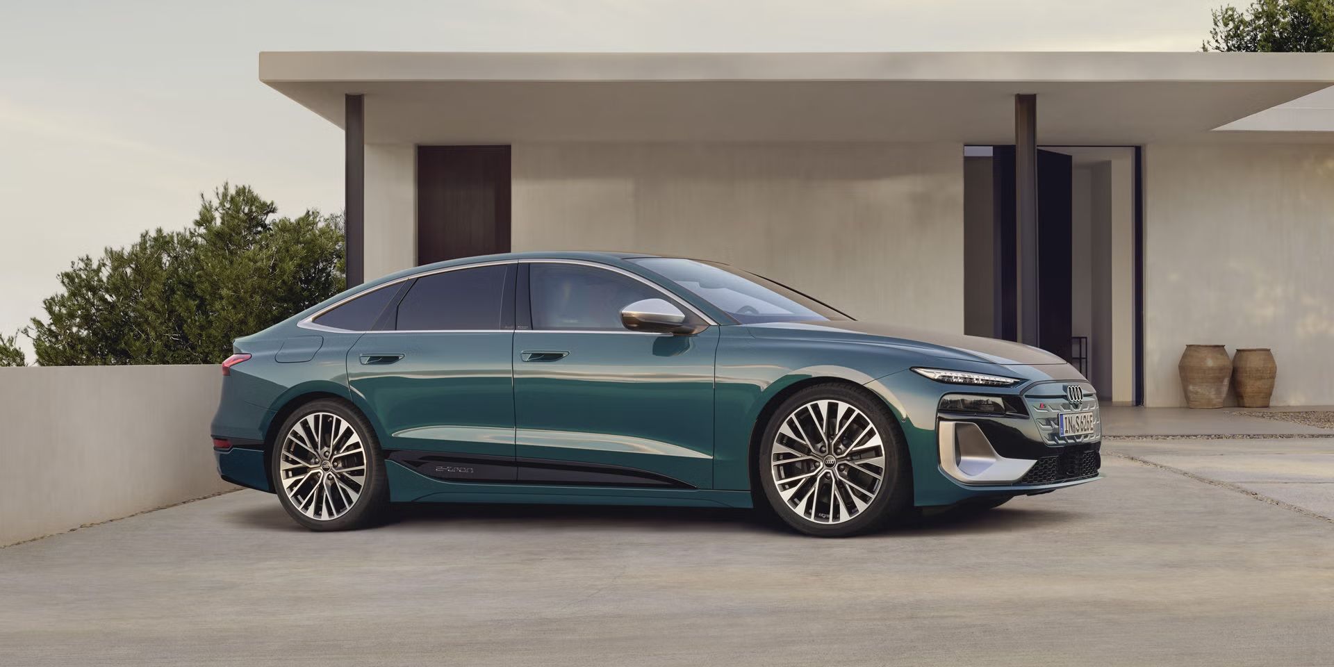 Električni Audi S6 e-tron je parkiran pred sodobno hišo in prikazan s strani. Vozilo stoji na urejenem dovozu v mirnem okolju. Poudarja čist dizajn in električno mobilnost znamke Audi.