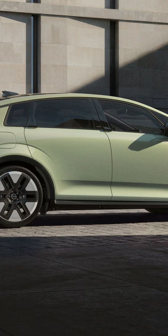 [AI-modified] – Audi A2 e-tron, fotografiran s strani, je parkiran pred sodobno stavbo v urbanem okolju. Poševna sončna svetloba delno potopi pokrajino v sence.