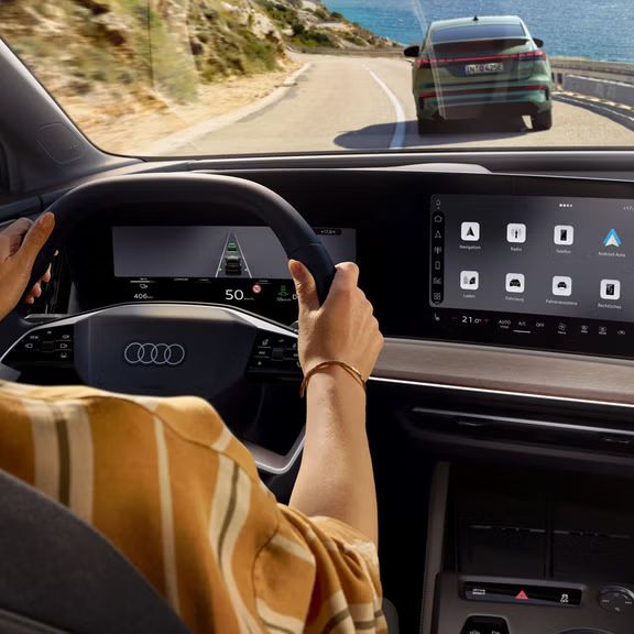 Pogled od zadaj na voznikov prostor modela Audi Q4 Sportback e-tron. Ženska sedi za volanom in vozi po gorski cesti, na desni strani je voda. Pred modelom Audi Q4 Sportback e-tron vozi drugo vozilo.