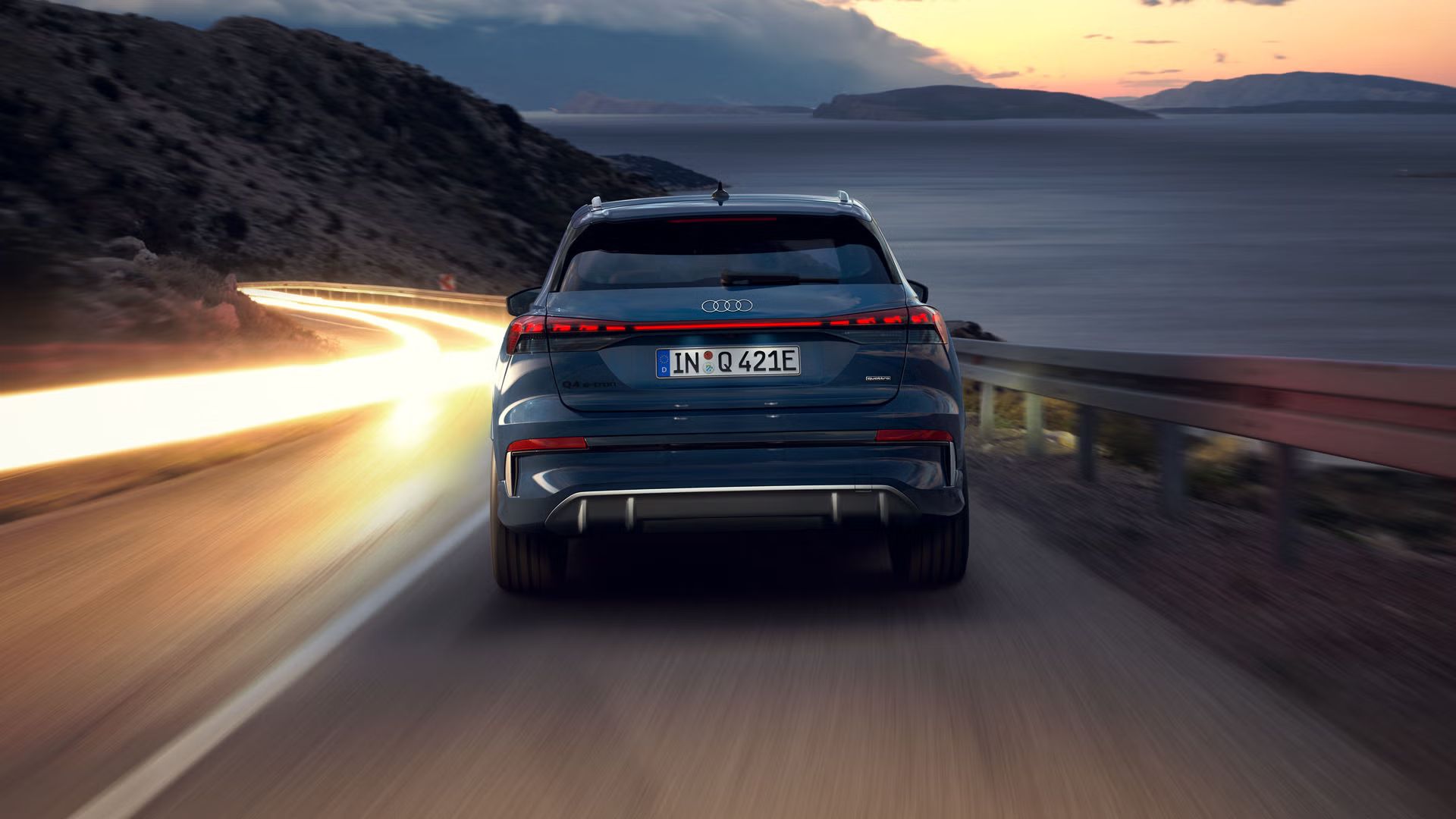 Der Q4 SUV e-tron von hinten bei Nacht auf einer abschüssigen Gebirgstraße, zur rechten das Meer. Auf der Gegenfahrbahn sind Lichtschleier entgegenkommender Fahrzeuge zu sehen. 