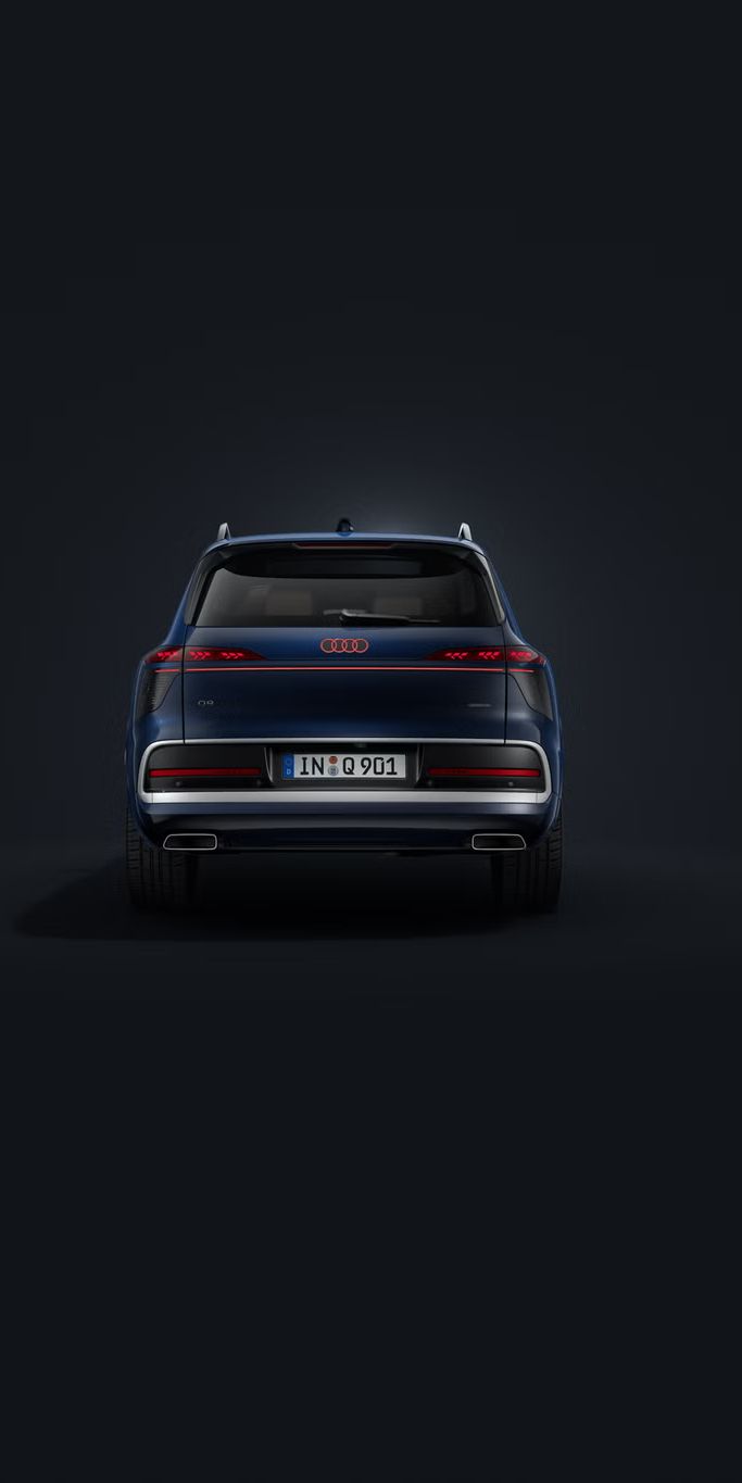Temno moder Audi Q9 SUV, fotografiran od zadaj na temnem studijskem ozadju, z neprekinjeno LED-svetlobno letvijo, osvetljenimi logotipi Audi in široko, markantno zadnjo zasnovo.