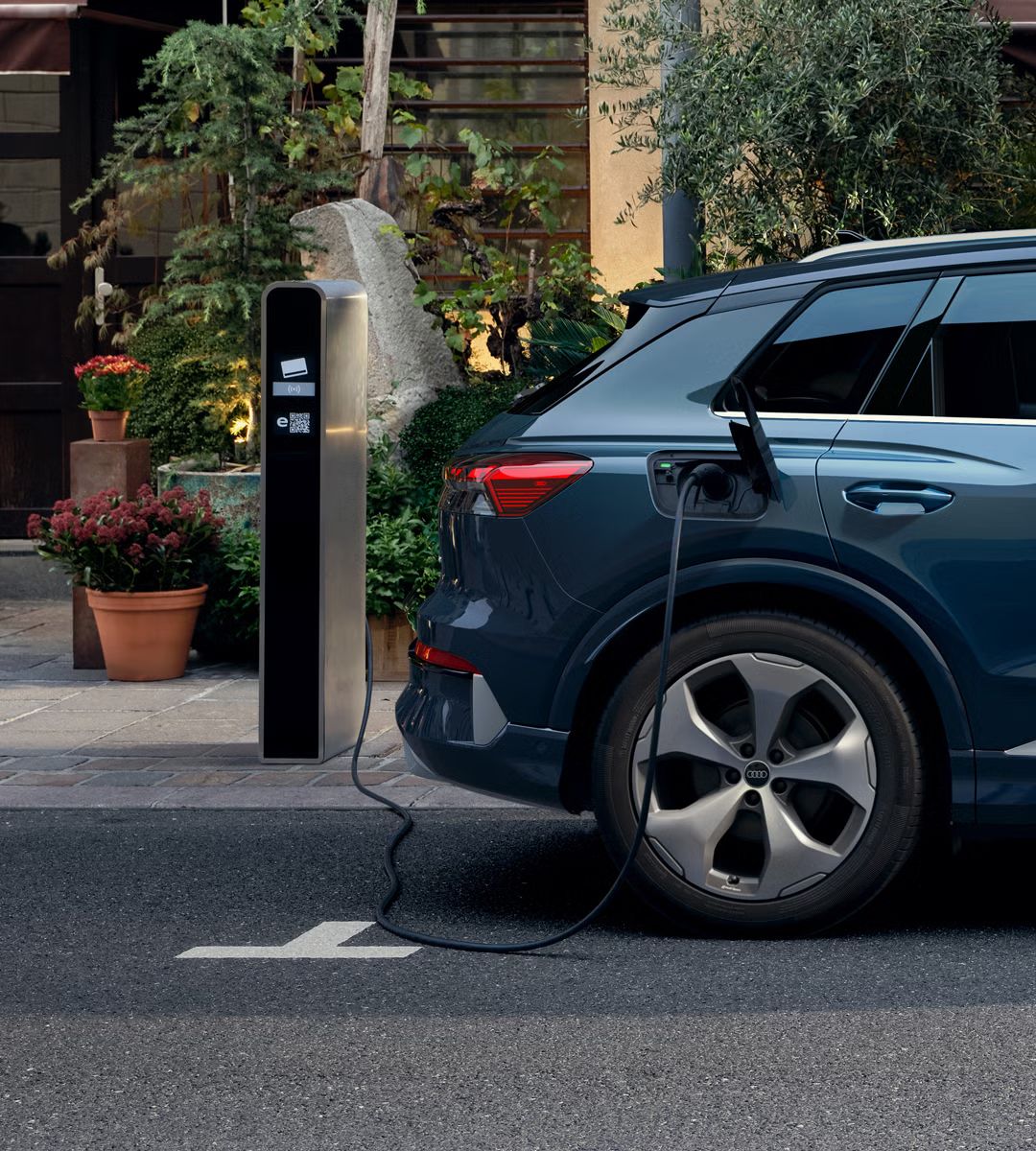 Bližnji posnetek desne strani zadnjega dela vozila Audi Q4 SUV e-tron, parkiranega pred stavbo. Pred stavbo so lonci z rožami. Vozilo je priključeno na javno polnilno postajo. 