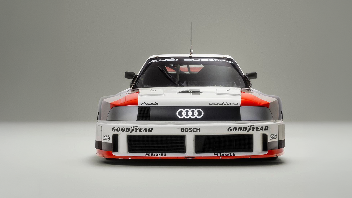 Dirkalni avtomobil Audi Quattro je fotografiran od spredaj na nevtralnem sivem ozadju. Avto ima široko, nizko karoserijo z izrazitim prednjim spojlerjem, logotipom Audi na sredini in vidnimi sponzorskimi napisi, kot sta Goodyear in Bosch. Luči so ozke in razpotegnjene, kar daje vozilu agresiven, športen videz.