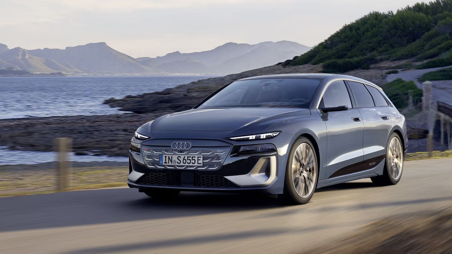 Električni Audi S6 e-tron vozi po obalni cesti ob morju, z griči v ozadju. Vozilo je prikazano s sprednje strani med vožnjo v naravnem okolju. Fotografija poudarja sodoben dizajn in električno mobilnost znamke Audi.