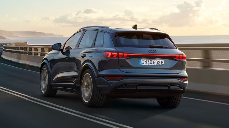 Poševni zadnji posnetek modela Audi Q6 SUV e-tron med vožnjo po vijugasti obalni cesti. V ozadju se dvigajo gore.