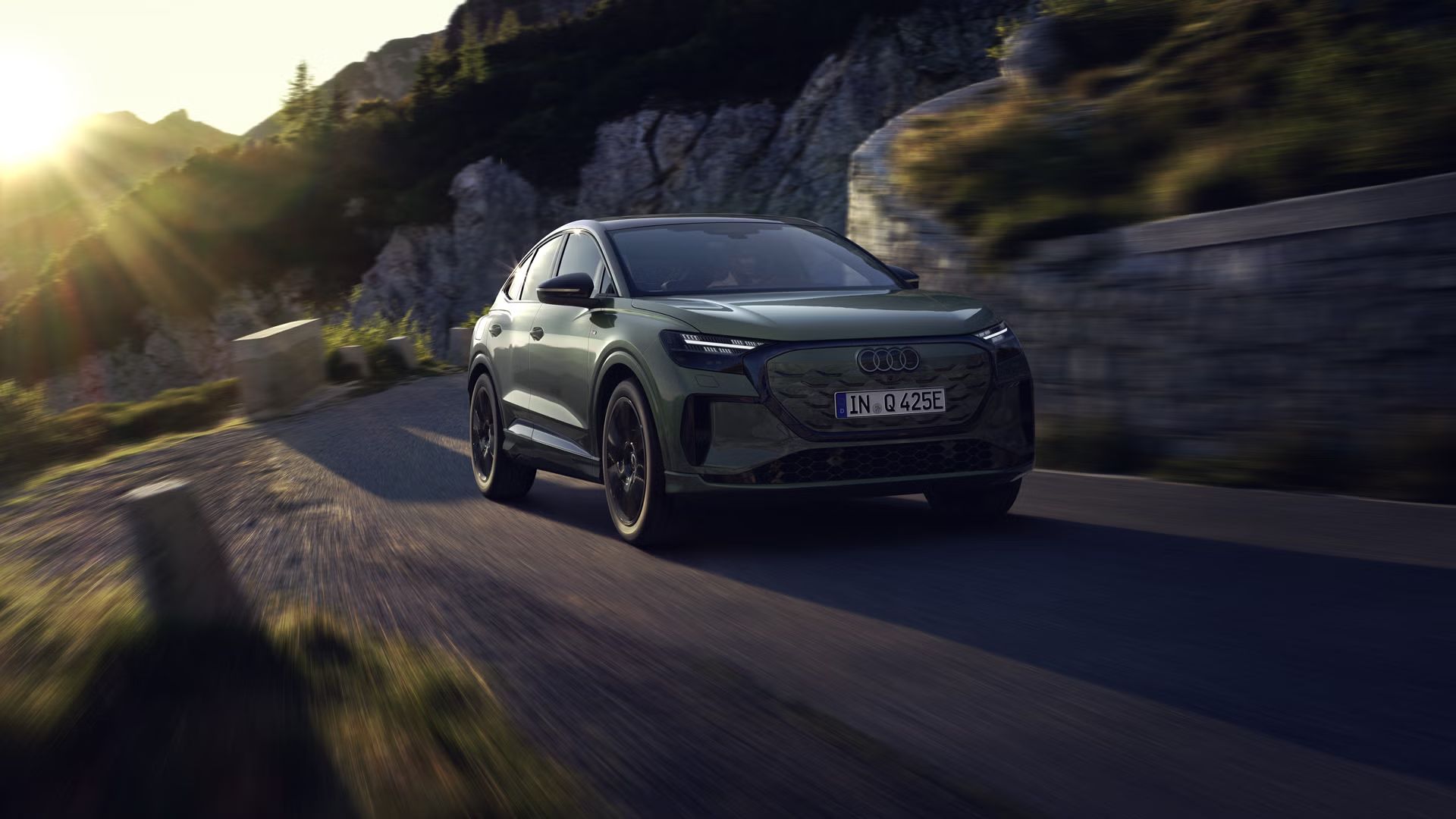 Audi Q4 Sportback e-tron med vožnjo po gorski cesti. Sliko z leve zgornje strani osvetljujejo sončni žarki, vozilo meče senco predse.