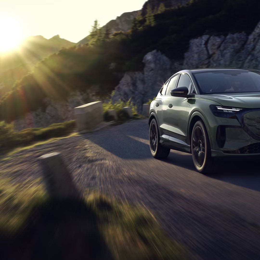 Audi Q4 Sportback e-tron med vožnjo po gorski cesti. Sliko z leve zgornje strani osvetljujejo sončni žarki, vozilo meče senco predse.