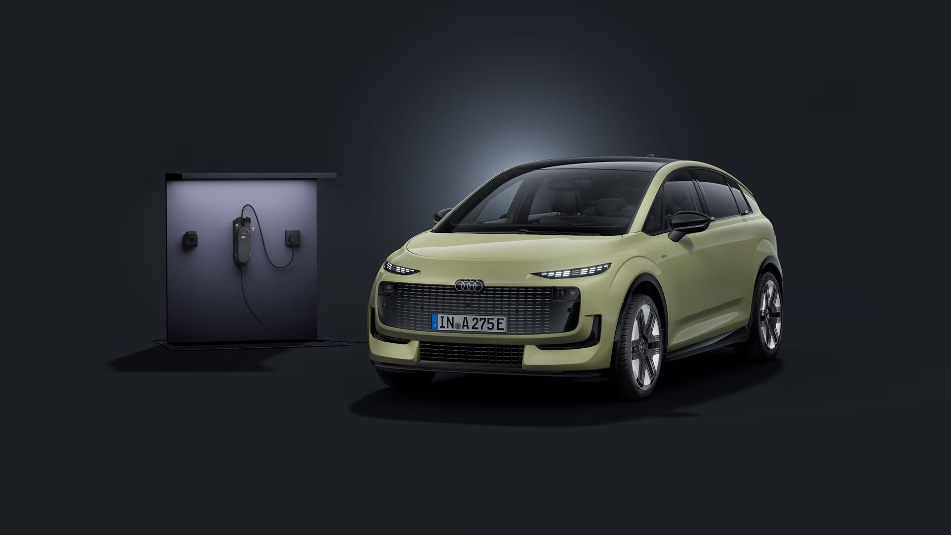 Stranski sprednji del vozila Audi A2 e-tron s temnim ozadjem, levo za njim je polnilna postaja.