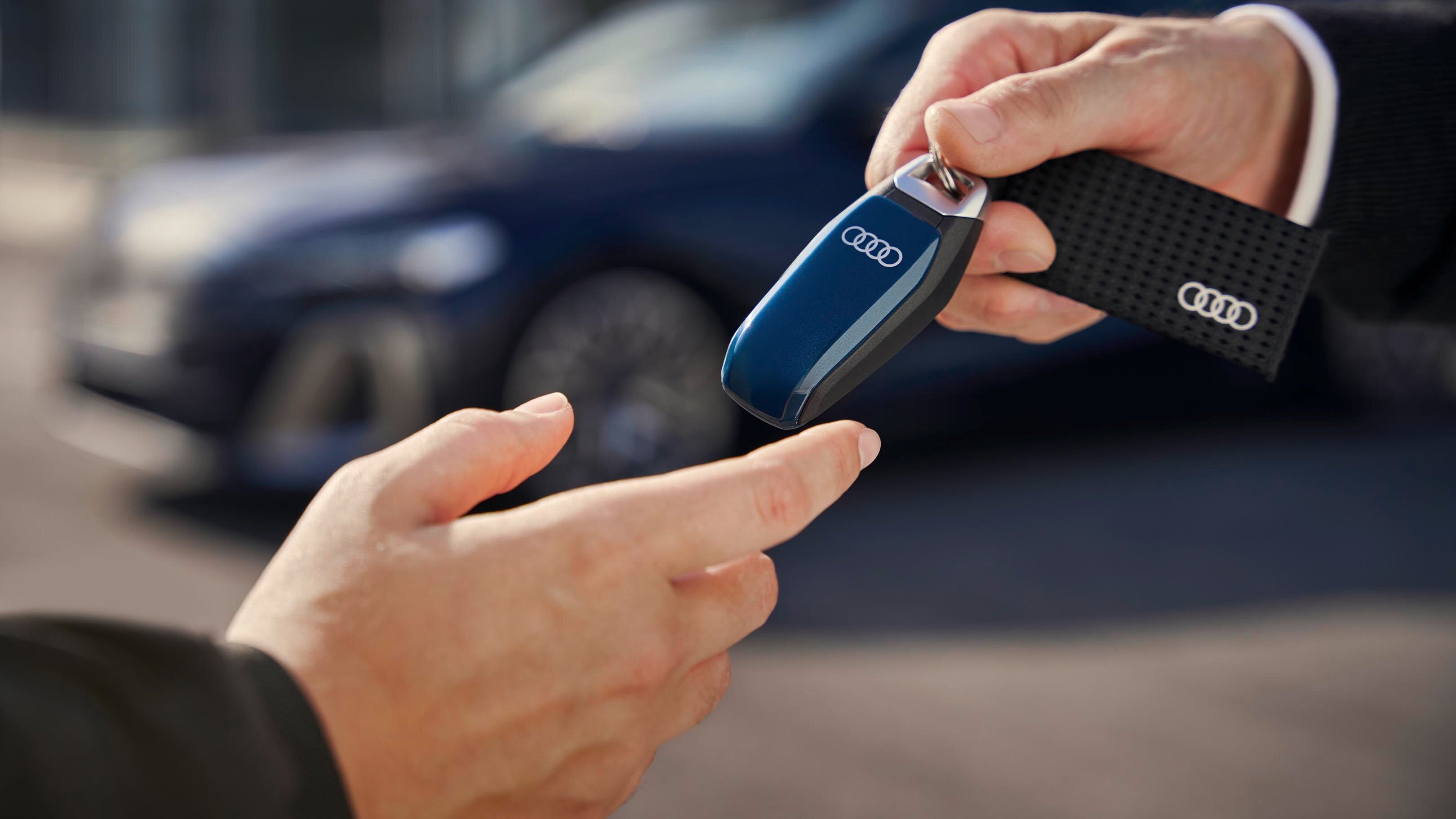 Oseba izroča ključe avtomobila drugi osebi. V ozadju je parkiran avto znamke Audi. Izmenjava poteka na prostem.