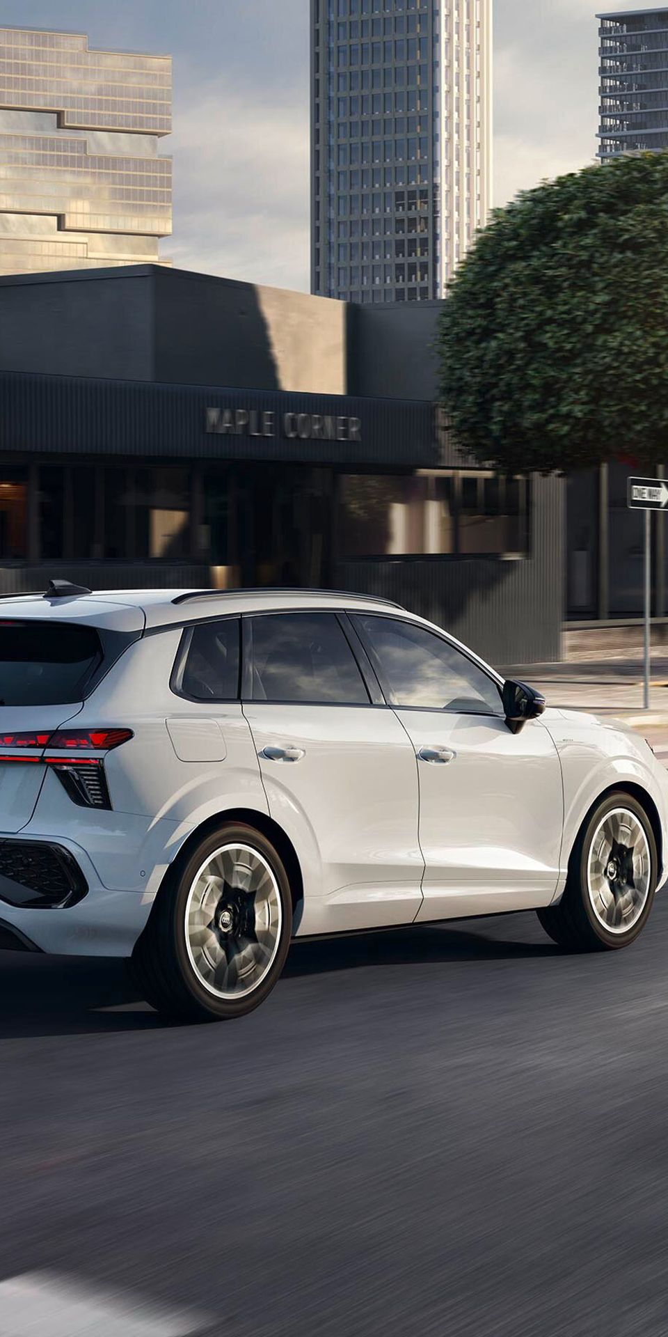 Audi Q3 SUV e-hybridi v beli barvi vozi po mestni ulici s sodobnimi zgradbami v ozadju.