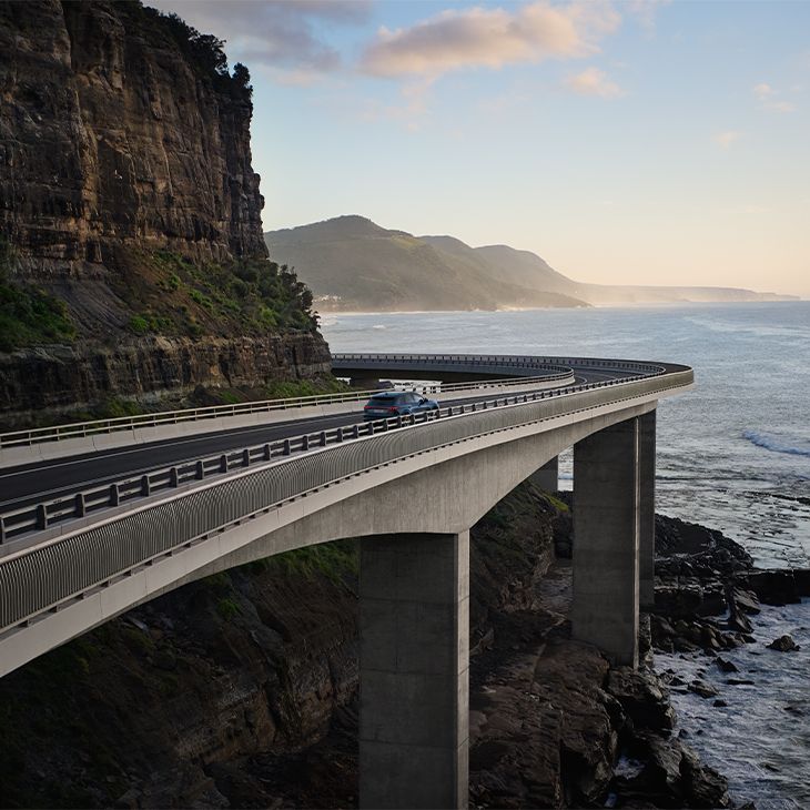 Dolga, ukrivljena cesta na betonskih stebrih poteka ob strmem skalnatem klifu nad oceanskimi valovi. Avto vozi po viaduktu, obdanem z zaščitno ograjo, medtem ko se v ozadju vidijo megličasti hribi in jutranja ali večerna svetloba.