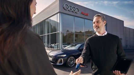 Prodajalec pred salonom Audi izroča ključe avtomobila stranki, ki stoji nasproti njega. V ozadju je viden moder Audi parkiran pred sodobno stekleno stavbo z logotipom znamke. Prodajalec v drugi roki drži tablico.